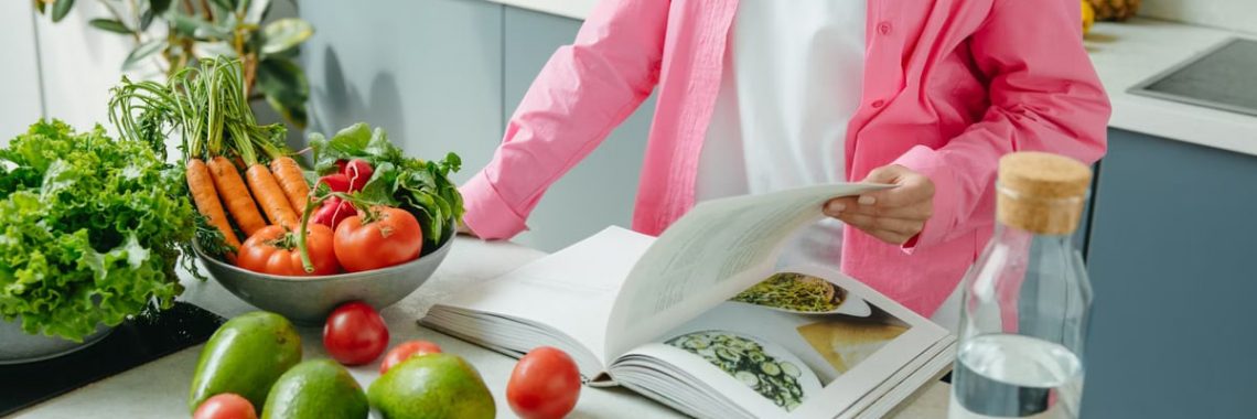 A person browsing a cookbook surrounded by fresh vegetables in a bright kitchen setting.